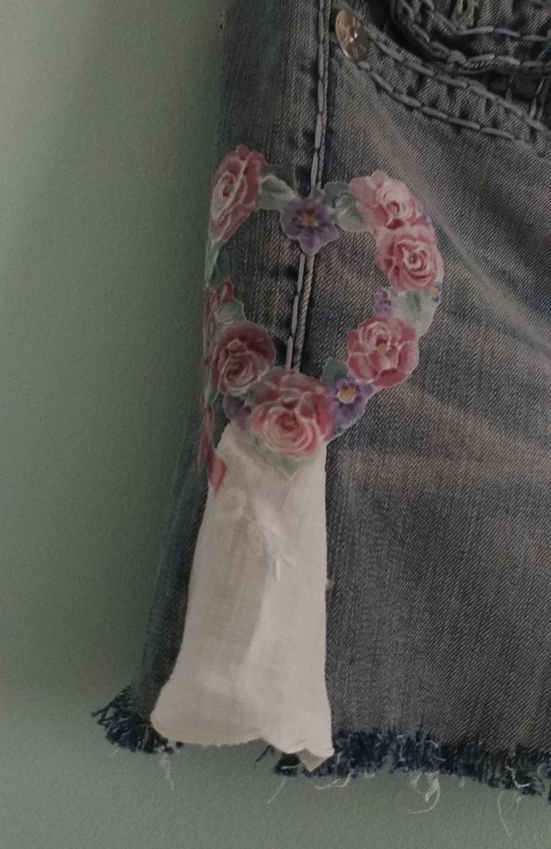 May include: A pair of denim shorts with a floral heart design on the front. The heart is made of pink, purple, and green flowers. The shorts have a frayed hem.