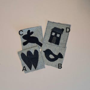 Puede incluir: Cuatro cuadrados de tela vaquera con dise&ntilde;os aplicados en negro: una letra "W", un p&aacute;jaro, un conejo y una casa. Los cuadrados est&aacute;n dispuestos sobre una superficie rosa claro. Los dise&ntilde;os est&aacute;n cosidos sobre la tela vaquera.