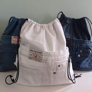 May include: Three drawstring bags made from repurposed denim jeans. One white bag and two blue denim bags are visible. The white bag has a "True Religion" label. Each bag has a black drawstring closure.