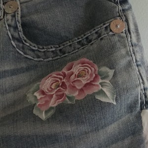 May include: Close-up of a pair of blue denim jeans with a floral design. The jeans have a pocket with a metal button and a decorative stitching detail. The floral design features two pink roses with green leaves.