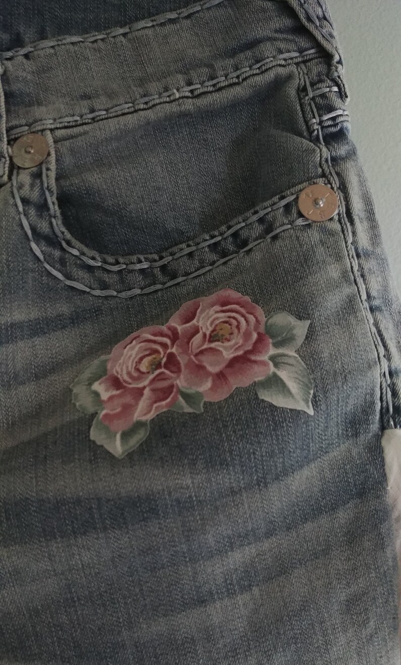 May include: Close-up of a pair of blue denim jeans with a floral design. The jeans have a pocket with a metal button and a decorative stitching detail. The floral design features two pink roses with green leaves.