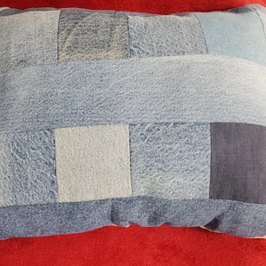 Pode incluir: Almofada retangular de ganga com um design de patchwork. A almofada apresenta quadrados e retângulos de ganga azul em várias tonalidades, criando um padrão texturizado e visualmente interessante. A almofada está colocada numa superfície vermelha.