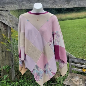 May include: A patchwork poncho in shades of pink, beige, and purple, with floral print accents. The poncho has a round neckline with a magenta trim and is adorned with small blue buttons. The garment is displayed outdoors.