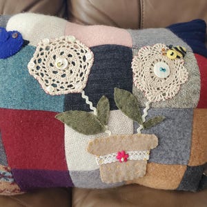 May include: A patchwork pillow with a floral design. The pillow features a blue bird and a felt flower pot with crocheted flowers and leaves. The pillow is made of various fabrics in shades of gray, blue, red, and beige.