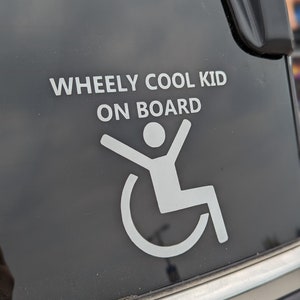 May include: A white sticker on a black car window that says "Wheely Cool Kid On Board" with a white silhouette of a person in a wheelchair.