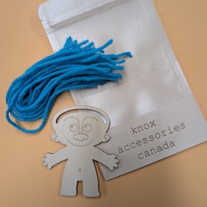 May include: A wooden doll with a blue yarn hairpiece. The doll is on a brown surface with a brown paper bag with the text "knox accessories canada".