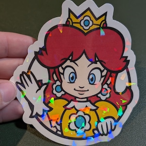 Peut inclure: Autocollant holographique représentant un personnage de dessin animé avec des cheveux roux, une robe jaune et une couronne. Le personnage fait un signe de la main. Bordure blanche et finition irisée réfléchissante.
