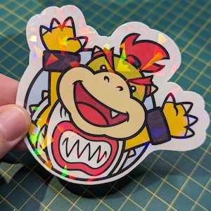 May include: A holographic sticker featuring a cartoon character with a red mohawk, yellow shell, and a toothy grin. The character is wearing a blue and black armband and is waving with one hand.