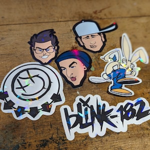 Op de afbeelding: Een set van vijf stickers met de band Blink-182. De stickers bevatten een logo met een smiley en pijlen, een konijn dat een blauwe spijkerbroek draagt en drie cartoon gezichten van de bandleden.