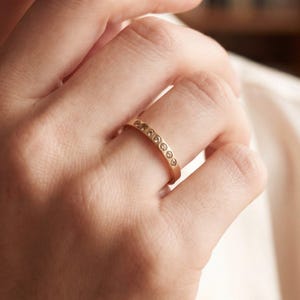 Può includere: Un anello d'oro con una fila di piccole pietre preziose rotonde e trasparenti. L'anello è su un dito, e la mano è in primo piano. L'anello è una semplice fascia con le pietre preziose disposte in fila. L'oro ha una finitura calda e lucida.
