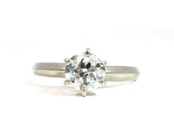 Op de afbeelding: Een zilverkleurige metalen ring met een enkele ronde, gefacetteerde heldere edelsteen in een zetting met zes pootjes. De ringband is glad en effen, met een gepolijste afwerking die het licht reflecteert over het ronde oppervlak en de steen.