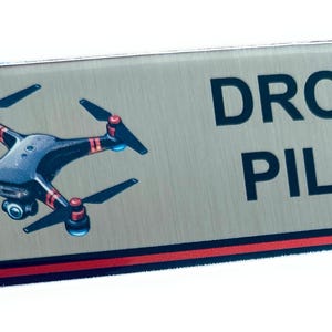 May include: A rectangular name tag with a brushed metal finish. The name tag features a detailed illustration of a drone in black and red, with the words "DRONE PILOT" in bold, dark blue letters. A thin red stripe accents the bottom.