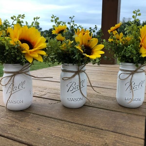 May include: Three white painted mason jars with yellow sunflowers and green foliage. Each jar is tied with twine and has the word "Ball" and "MASON" printed on the front. The jars are arranged on a wooden table.