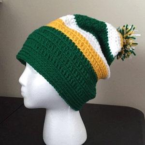 May include: A handmade crochet beanie in green, white, and yellow. The hat features a striped pattern and a matching pom-pom. The beanie is displayed on a white mannequin head, showcasing its shape and design. This winter hat is perfect for cold weather.
