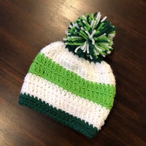 May include: A handmade crochet beanie hat with green, white, and dark green stripes, and a large pom-pom. The hat features alternating stripes of white and green, with a dark green brim. The pom-pom is made from green and white yarn.