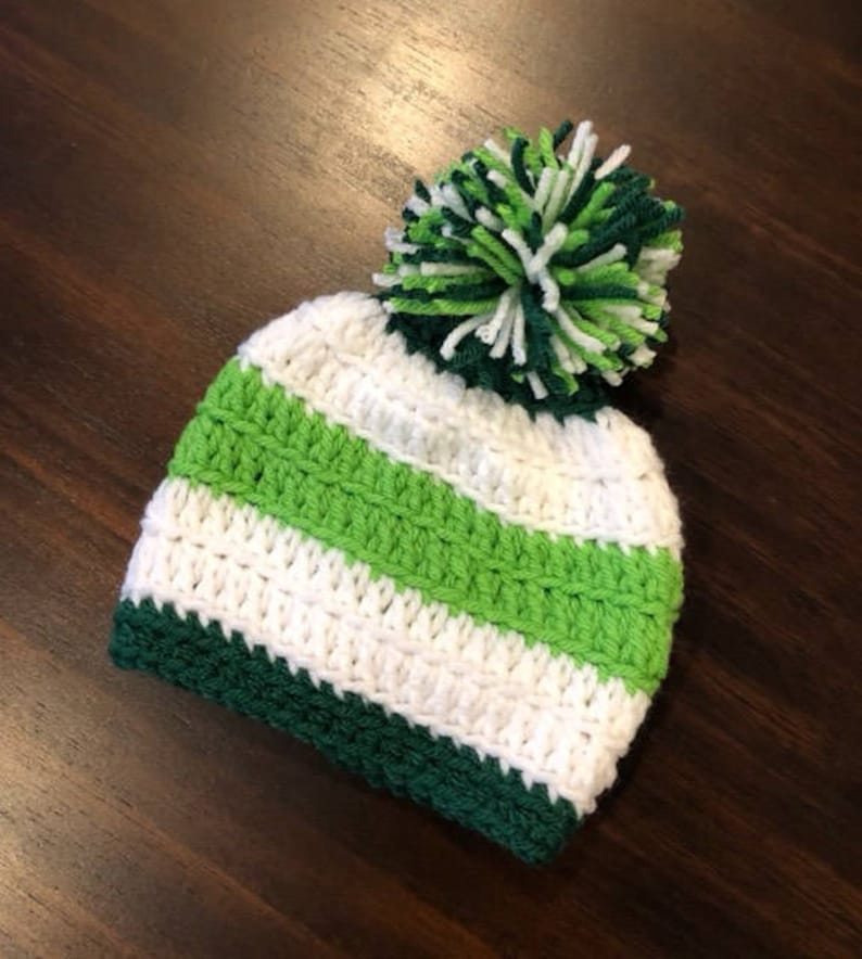 May include: A handmade crochet beanie hat with green, white, and dark green stripes, and a large pom-pom. The hat features alternating stripes of white and green, with a dark green brim. The pom-pom is made from green and white yarn.
