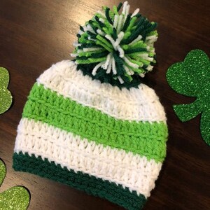 May include: A hand-crocheted hat with a green, white, and dark green striped design. The hat has a large pom-pom on top made of green and white yarn. This hat is likely for a baby or child, and is ideal for St. Patrick's Day celebrations.