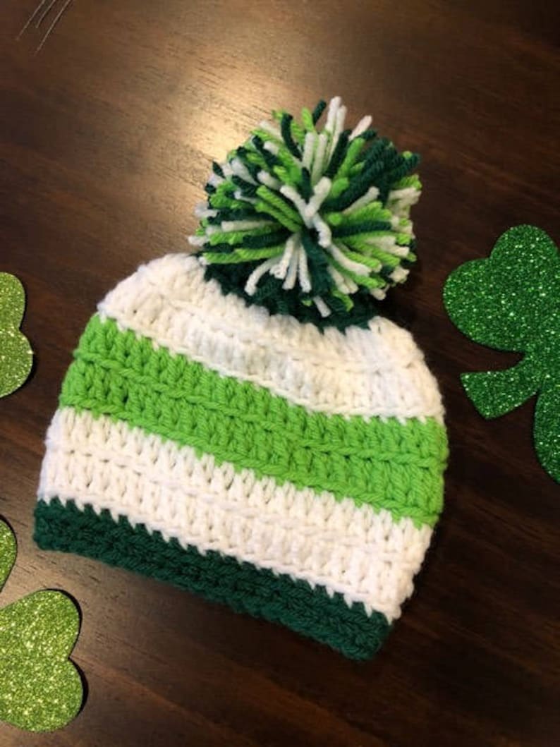 May include: A hand-crocheted hat with a green, white, and dark green striped design. The hat has a large pom-pom on top made of green and white yarn. This hat is likely for a baby or child, and is ideal for St. Patrick's Day celebrations.