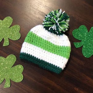 May include: A crochet beanie in white, green, and dark green, with a green and white pom-pom. Three glittery green shamrock cutouts are arranged around the hat on a dark wood surface. A festive St. Patrick's Day theme.