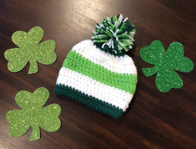 May include: A crochet beanie in white, green, and dark green, with a green and white pom-pom. Three glittery green shamrock cutouts are arranged around the hat on a dark wood surface. A festive St. Patrick's Day theme.