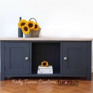 May include: A dark grey wooden cabinet with three doors and a shelf. The cabinet has a light wood top and is sitting on a wooden floor. There are sunflowers and a bottle on the top of the cabinet.