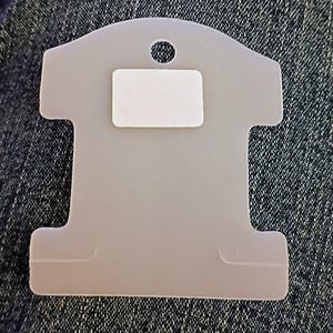 May include: A light gray, T-shaped jewelry display card with a small hole at the top and a rectangular adhesive pad. The card is designed to hold necklaces or other jewelry items. The background is a textured denim fabric.