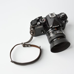 Può includere: Una macchina fotografica a pellicola Nikon nera con una tracolla in pelle marrone. La macchina fotografica ha un obiettivo con un parasole.