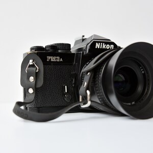 Peut inclure: Un appareil photo argentique Nikon FM3A noir avec un objectif et une sangle en cuir noir.