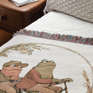 May include: A white woven throw blanket with a fringe border features a whimsical illustration of two frogs riding a bicycle. The frogs are wearing clothes and the bicycle has a basket.