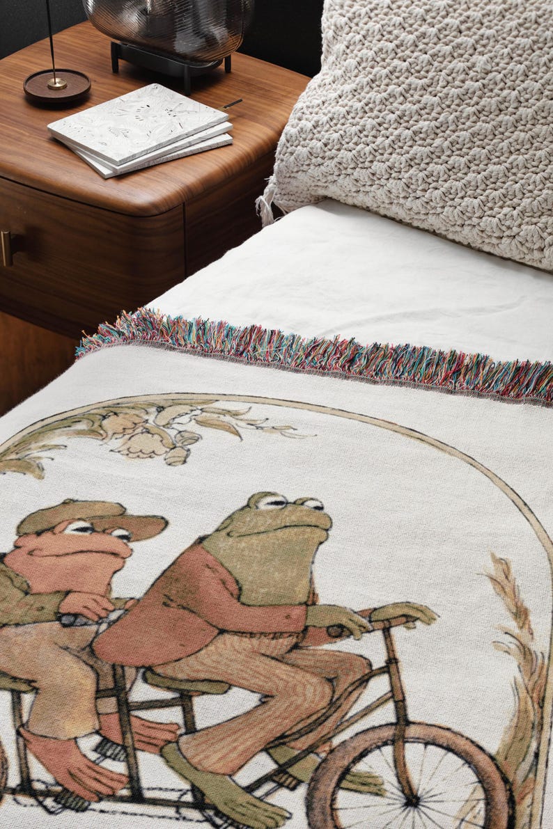 May include: A white woven throw blanket with a fringe border features a whimsical illustration of two frogs riding a bicycle. The frogs are wearing clothes and the bicycle has a basket.