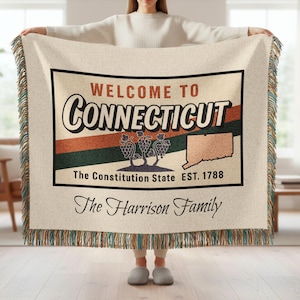 Könnte beinhalten: Cremefarbene gewebte Wurfadecke mit Fransen. Die Decke hat ein Design im Vintage-Stil mit der Aufschrift "Welcome to Connecticut" mit einer Karte des Staates, Trauben und dem Text "The Constitution State EST. 1788". Die Worte "The Harrison Family" stehen ebenfalls auf der Decke.