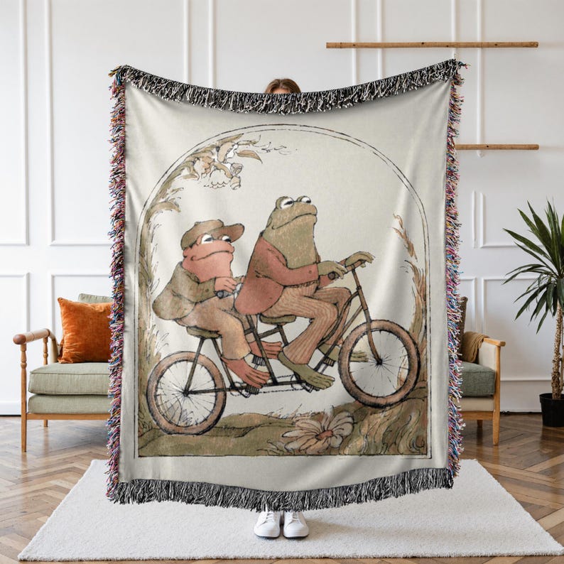 May include: Two frogs riding a bicycle together. The frog in the front is wearing a red jacket and the frog in the back is wearing a brown jacket. The bicycle is brown with a white seat and white handlebars.