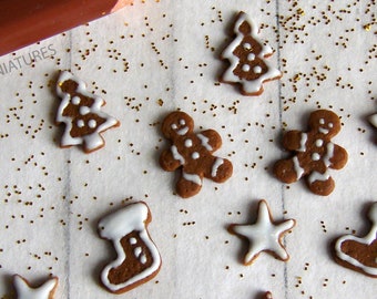 May include: Miniature gingerbread cookies in various shapes, including gingerbread men, Christmas trees, stars, and stockings, are arranged on a white surface with gold glitter. A brown pencil is in the upper left corner. The text "tinyartminiatures" is visible.