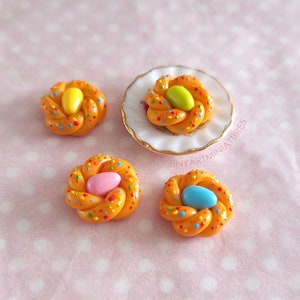 Pode incluir: Quatro miniaturas de pastelaria entran&ccedil;ada cor de laranja com granulado colorido e um grande doce no centro. Uma pastelaria est&aacute; sobre um prato branco pequeno com borda dourada. Os doces s&atilde;o amarelos, verdes, rosa e azuis.