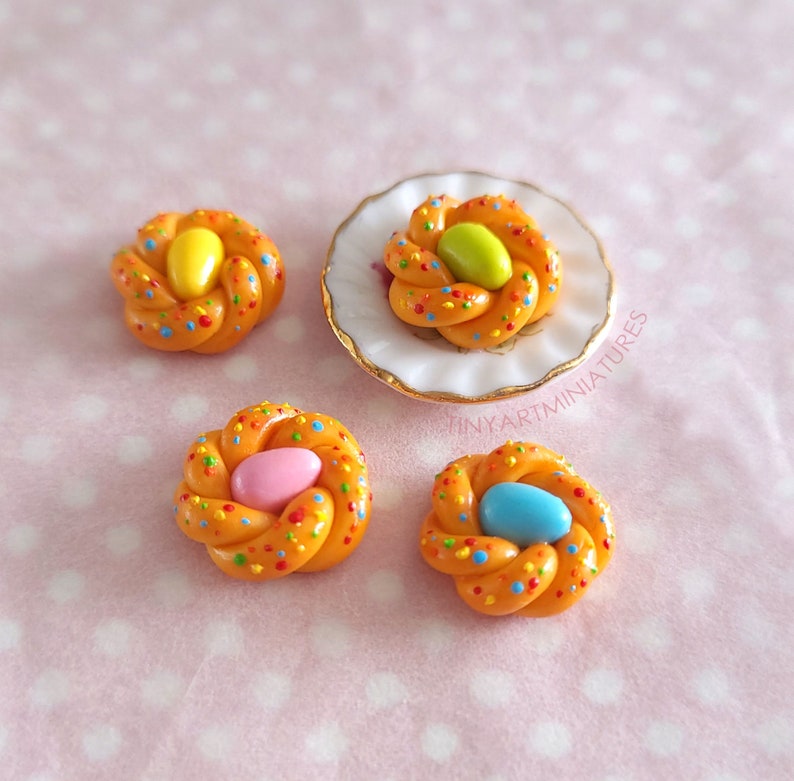 Pode incluir: Quatro miniaturas de pastelaria entran&ccedil;ada cor de laranja com granulado colorido e um grande doce no centro. Uma pastelaria est&aacute; sobre um prato branco pequeno com borda dourada. Os doces s&atilde;o amarelos, verdes, rosa e azuis.