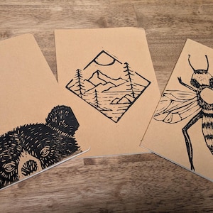 May include: Three kraft paper notebooks with black linocut designs. One features a bear's face, another a mountain landscape in a diamond, and the third a detailed bee illustration. The notebooks are arranged on a wooden surface.