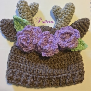 May include: A brown crocheted hat with purple flowers and green leaves, topped with two gray crocheted antlers. The hat has the word "Pattern" written on it.