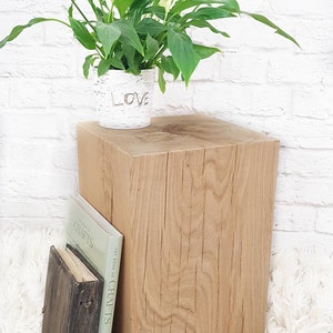 May include: A natural wood cube end table with a white ceramic planter with the word "LOVE" on it. The table is sitting on a white fluffy rug with a book titled "The Book of Crafts" on top of it. The table is made by All Things Oak Studio.