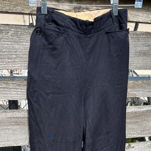 May include: A pair of solid black trousers featuring a high-waisted design with a button closure at the side, a structured waistband, and tapered legs that narrow toward the ankles, presented in a simple, clean, and functional cut.