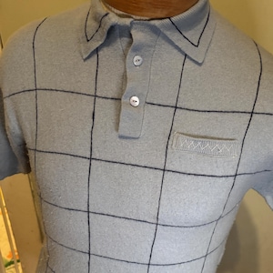Pode incluir: Camisa polo azul claro com um padrão de grade em azul marinho. A camisa tem gola, mangas curtas e um bolso no peito. A roupa é exibida em um manequim.