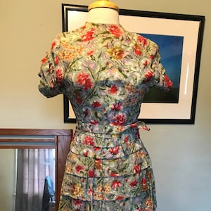 May include: A vintage floral print dress with a high neck, short sleeves, and a tiered skirt. The dress is made of a light blue fabric with a floral pattern in shades of red, pink, yellow, and green.