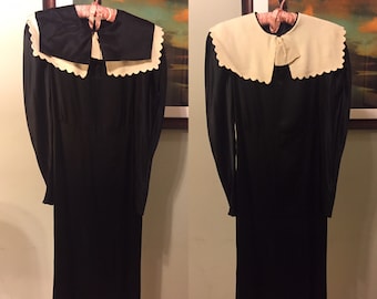 May include: A black satin dress with a large white collar and a bow at the neckline. The collar has a scalloped edge.