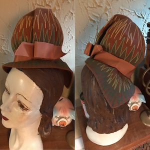 May include: A brown and green hat with a large, round crown and a wide brim. The hat has a brown ribbon tied in a bow at the front. The hat is sitting on a mannequin head.