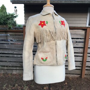 May include: A white suede jacket with fringe detailing and red and green embroidered flowers. The jacket has a zipper closure and two pockets.