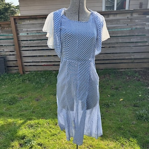 May include: A blue and white striped dress with short, white sleeves is displayed on a dress form. The dress has a diagonal stripe pattern and a flowing skirt. The dress form is on a metal stand, set against a backdrop of a wooden fence and green grass.