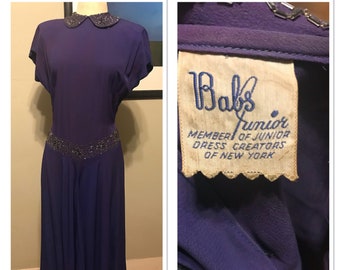 May include: A purple vintage dress with a white collar and beaded embellishment at the waist. The dress has a label that reads "Bab's Junior Member of Junior Dress Creators of New York."