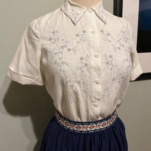 May include: A white short-sleeved button-down shirt with a floral embroidery design on the front. The shirt has a collar and a button closure. The shirt is paired with a navy blue skirt with a white and red embroidered waistband.