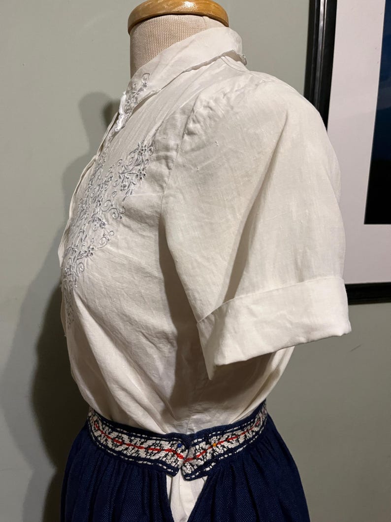 May include: A white embroidered short-sleeved blouse with a navy blue skirt with a white and red embroidered waistband.