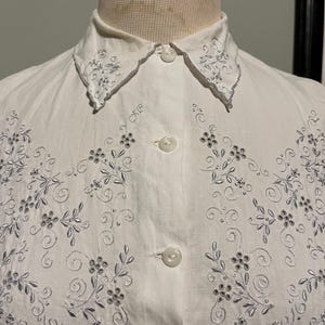 May include: White cotton button-up shirt with a delicate floral embroidery design in silver thread. The collar has a scalloped edge and is also embroidered.