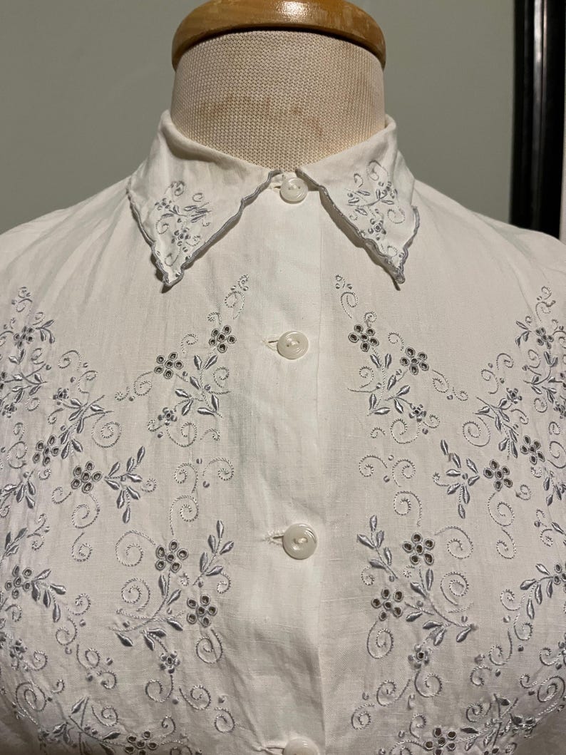 May include: White cotton button-up shirt with a delicate floral embroidery design in silver thread. The collar has a scalloped edge and is also embroidered.